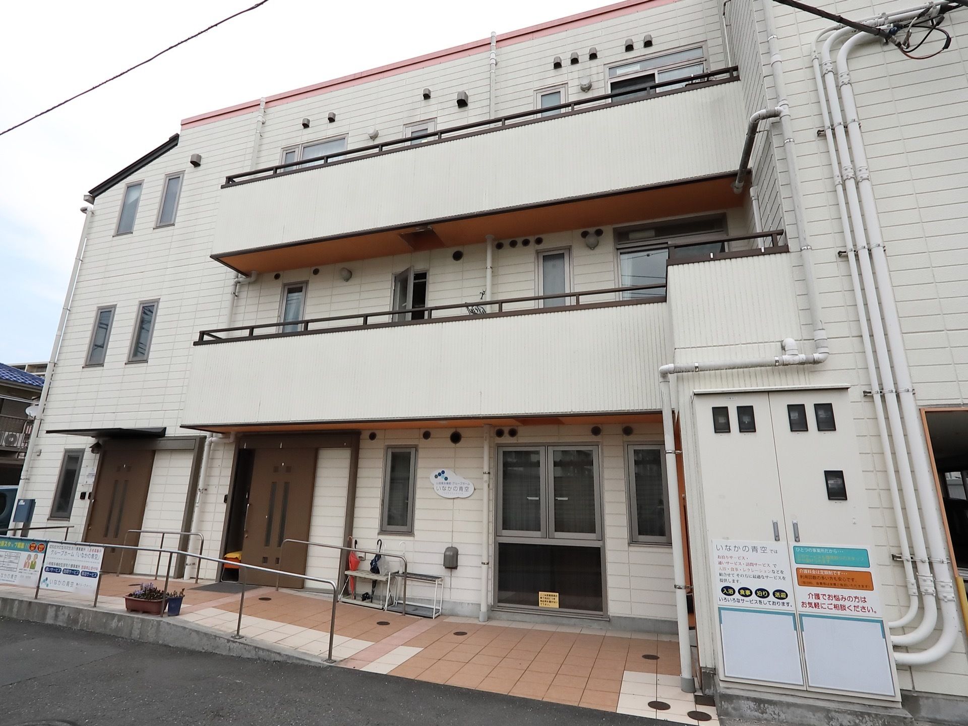 ３階建ての白い建物である。外壁にメーターボックスが取りつけられている。玄関扉は２か所にあり、傘立てが置かれている。