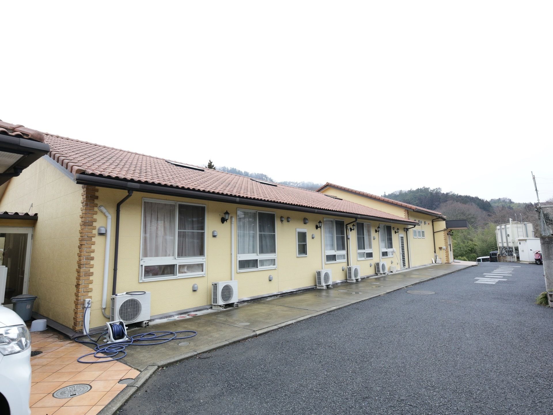 洋風の平屋建てで、明るい色合いの外壁の建物である。外にはエアコンの室外機が設置されており、散水用のホースがある。