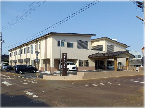 2階建ての建物は白い外壁に屋根が付き、窓もたくさんある。道路に面していて駐車スペースにはたくさんの車が止められている。