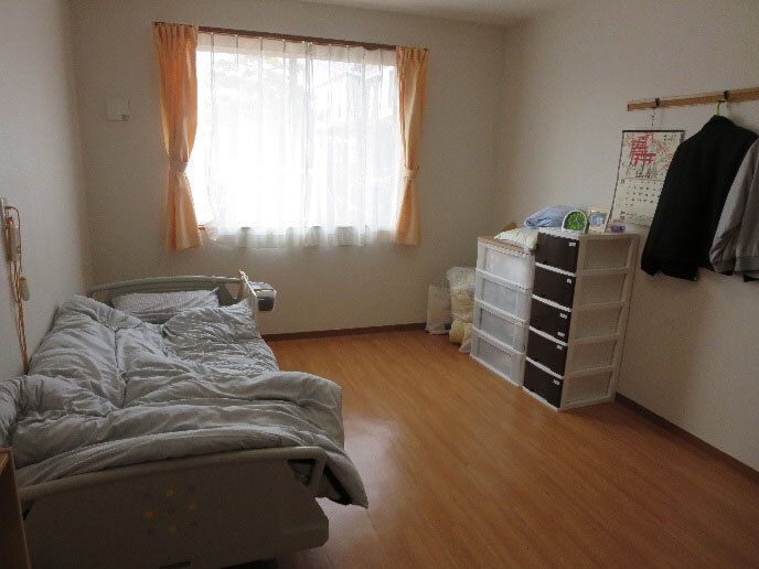 レースカーテンとカーテンが付いた大きな窓があるフローリングの部屋。ベッドサイドには緊急通報装置がある。