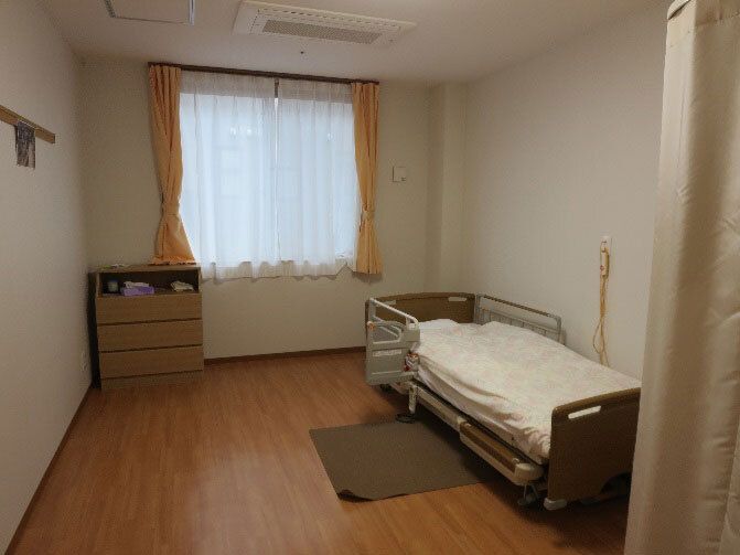 寝具付きのベッドが置かれたフローリングの部屋。ベッドサイドには緊急通報装置があり、窓にはレースカーテンとカーテンがある。