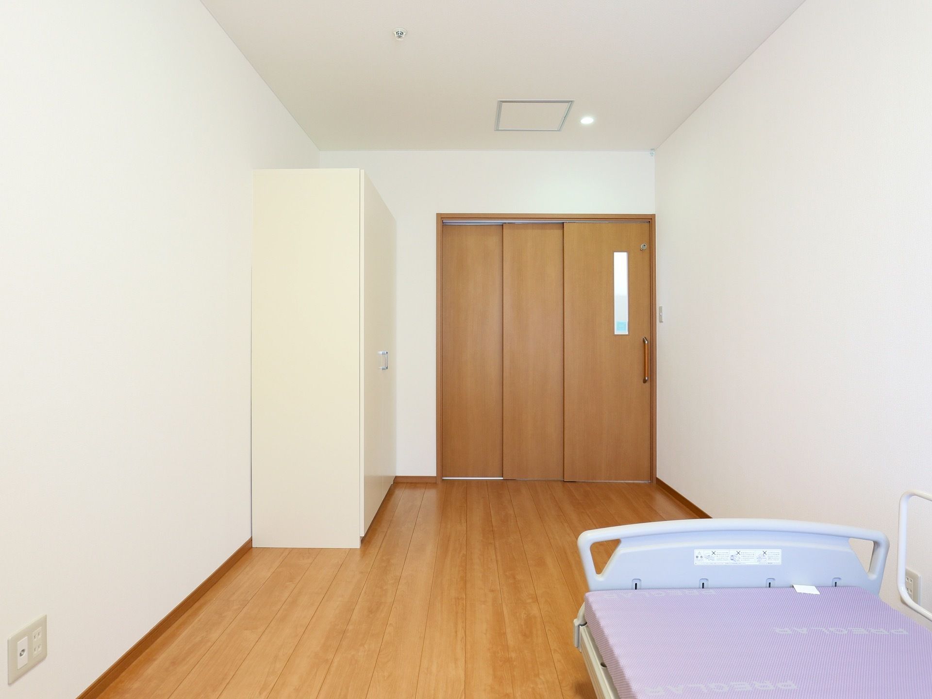 フローリングに白い壁で構成された居室である。右手前に介護用ベッドが置かれ、左奥に収納スペースが用意されている。