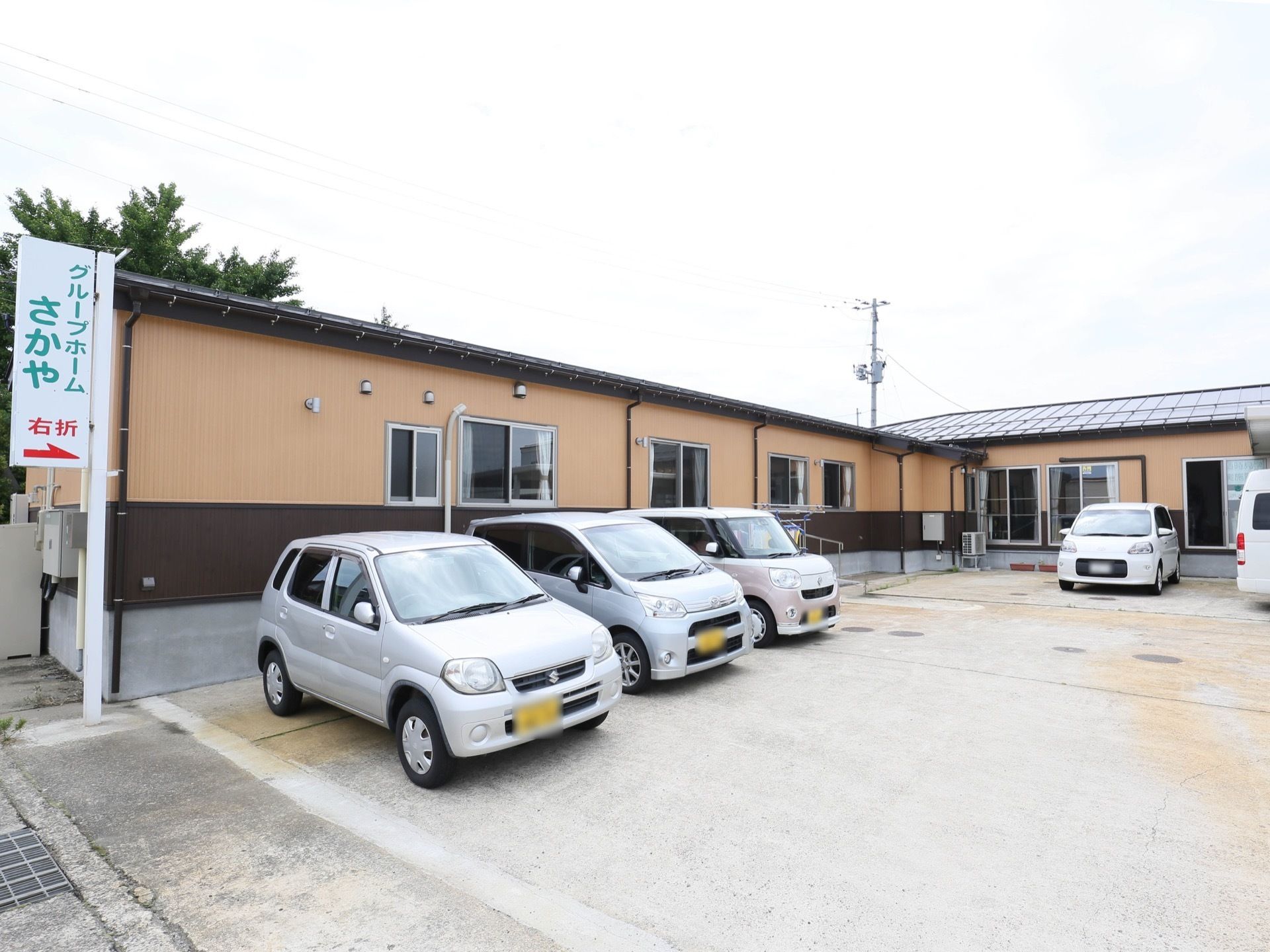 敷地内に平屋建ての建物があり、前には駐車場も完備されている。数台の車が停車しており、角には施設看板が設置されている。