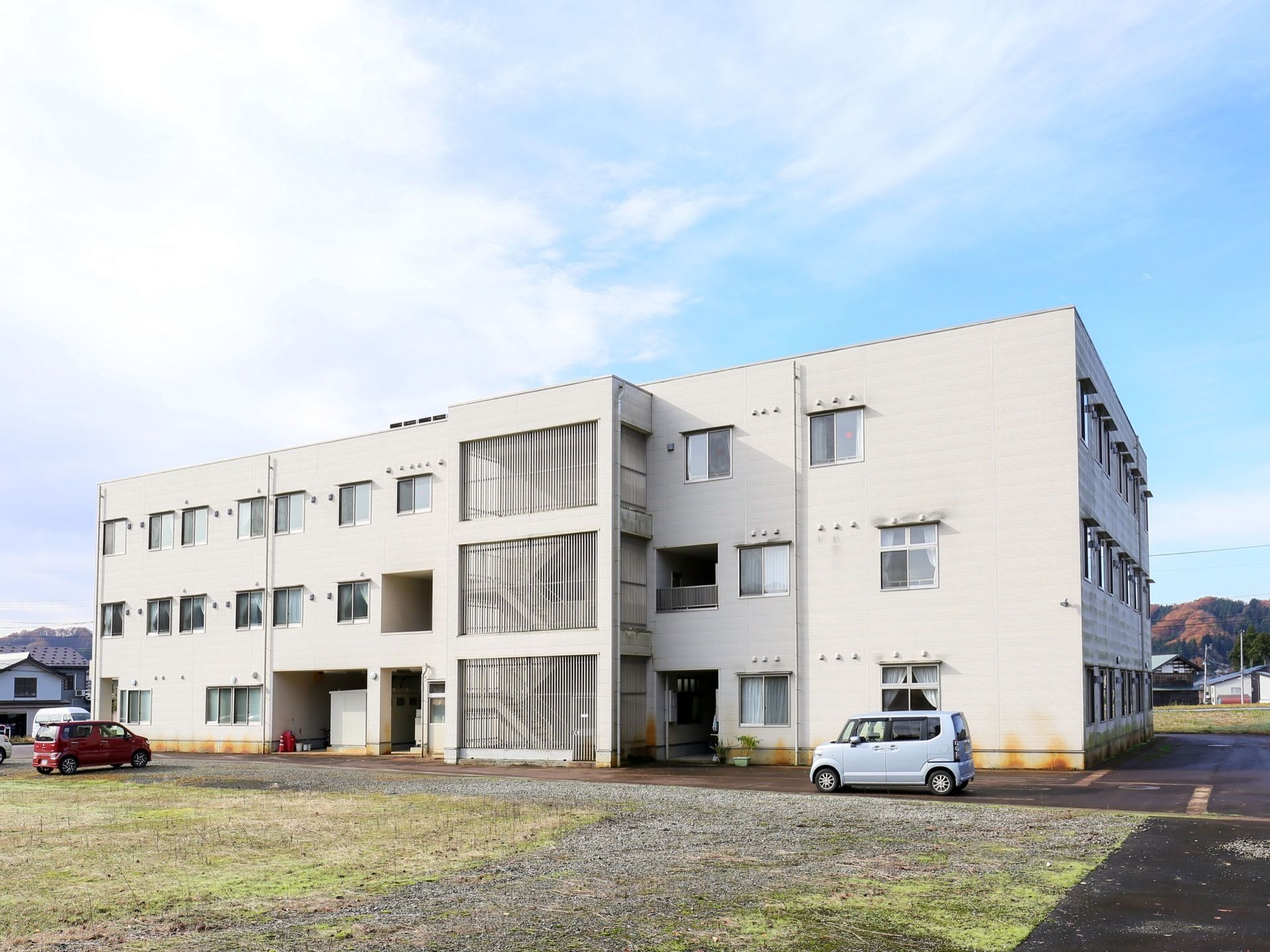 施設建物の裏側の様子である。手前に駐車場があり、奥に白い３階建ての建物がある。中央付近に、外階段が設けられている。