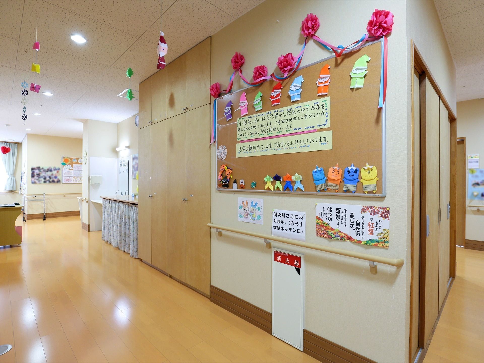 廊下の壁に、サンタクロースやキャラクターの姿をした紙細工などが貼られている。下の方に、消火器入れが設置されている。