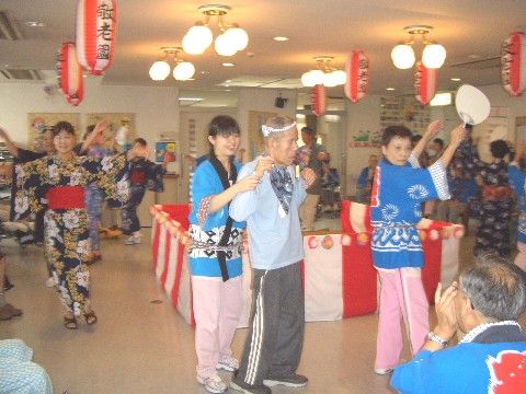室内で盆踊り大会が開催されている様子。提灯が飾られ、浴衣や法被姿で盆踊りが行われている。