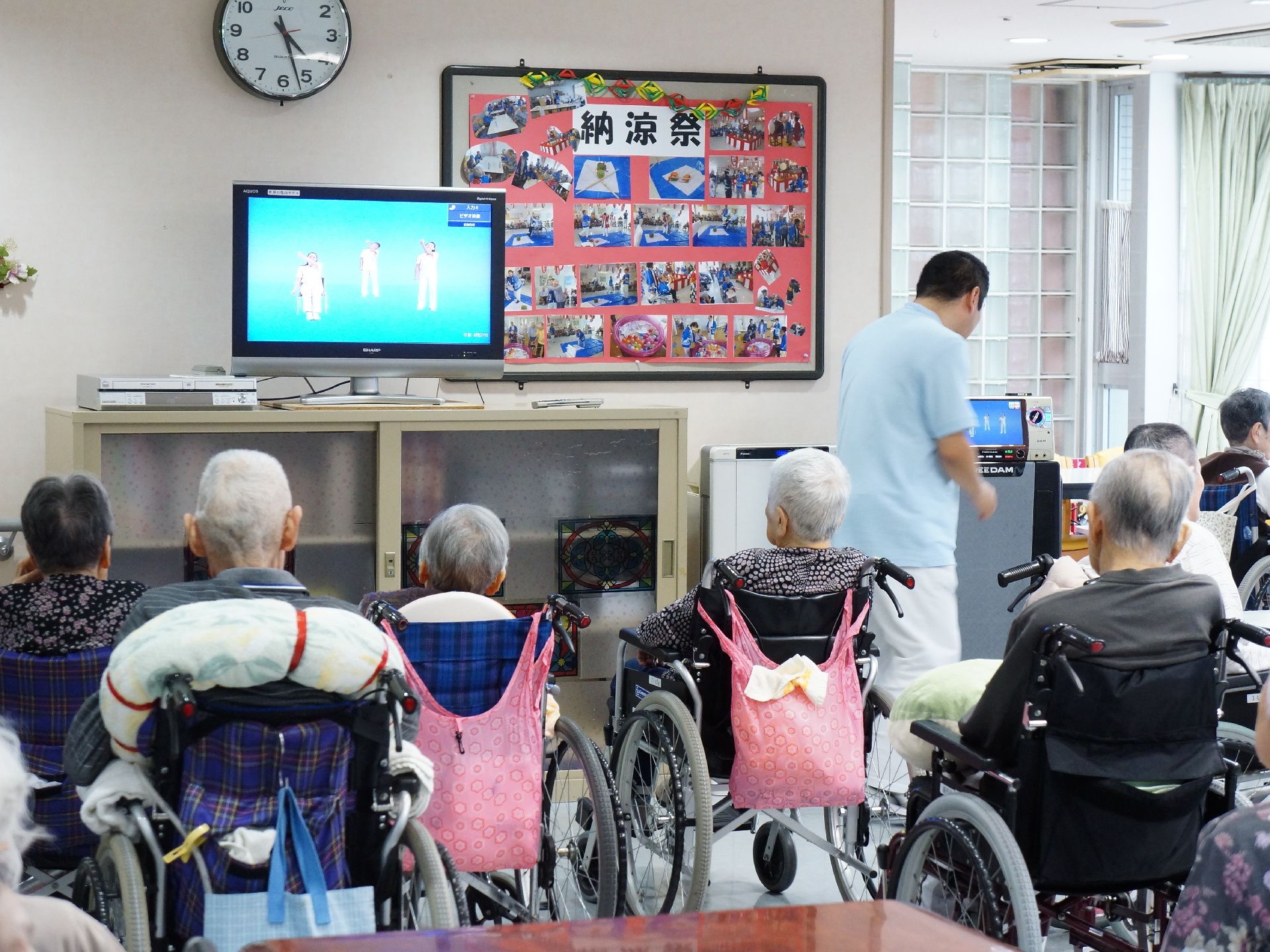 ホールに集まる利用者様の様子。テレビモニターには体操の映像が流れている。職員が準備をしている姿が見られる。
