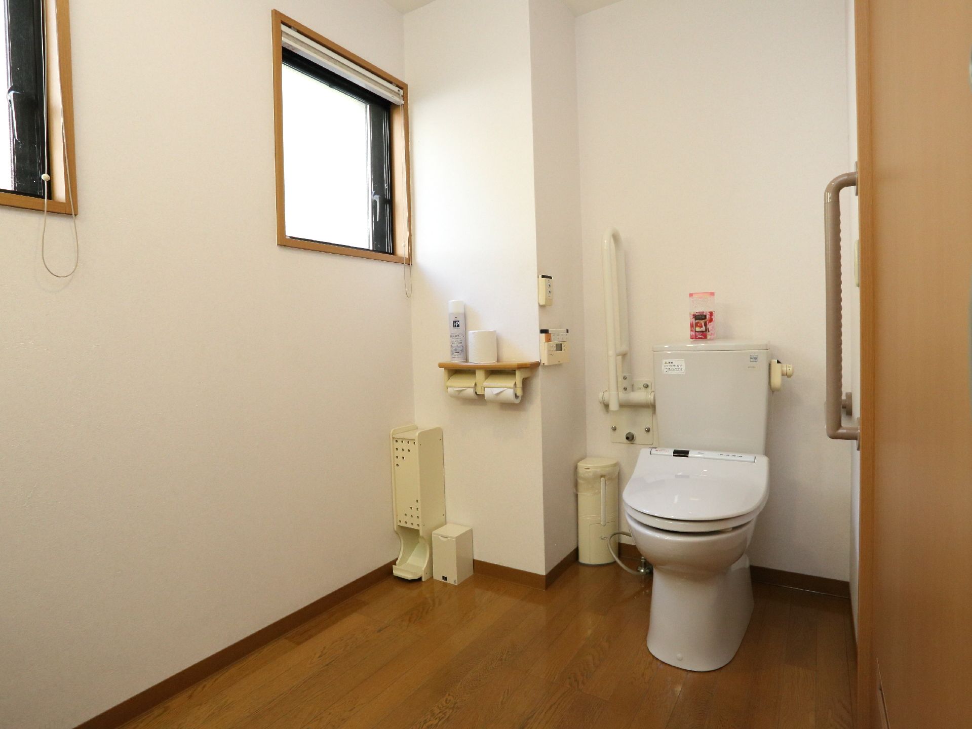 白色の壁に木目調の床のトイレである。横側には、四角形の小さな窓が並んでいる。便器の横側には、上げ下げの手すりを備えている。
