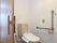 白色の壁に、木目調の床のトイレである。壁には、茶色のエル型の手すりとナースコール、トイレットペーパーホルダーを設置している。