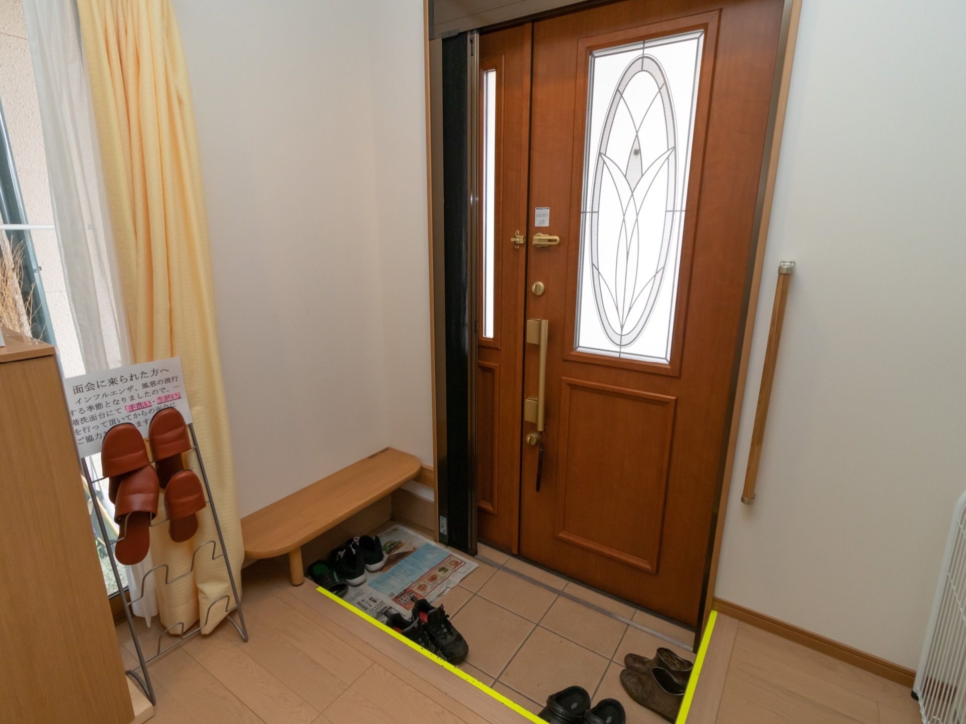 木目調の玄関ドアが設置されていて、ドアの一部にステンドグラス調のガラスがはめ込まれている。ドアの横の壁に手すりがある。
