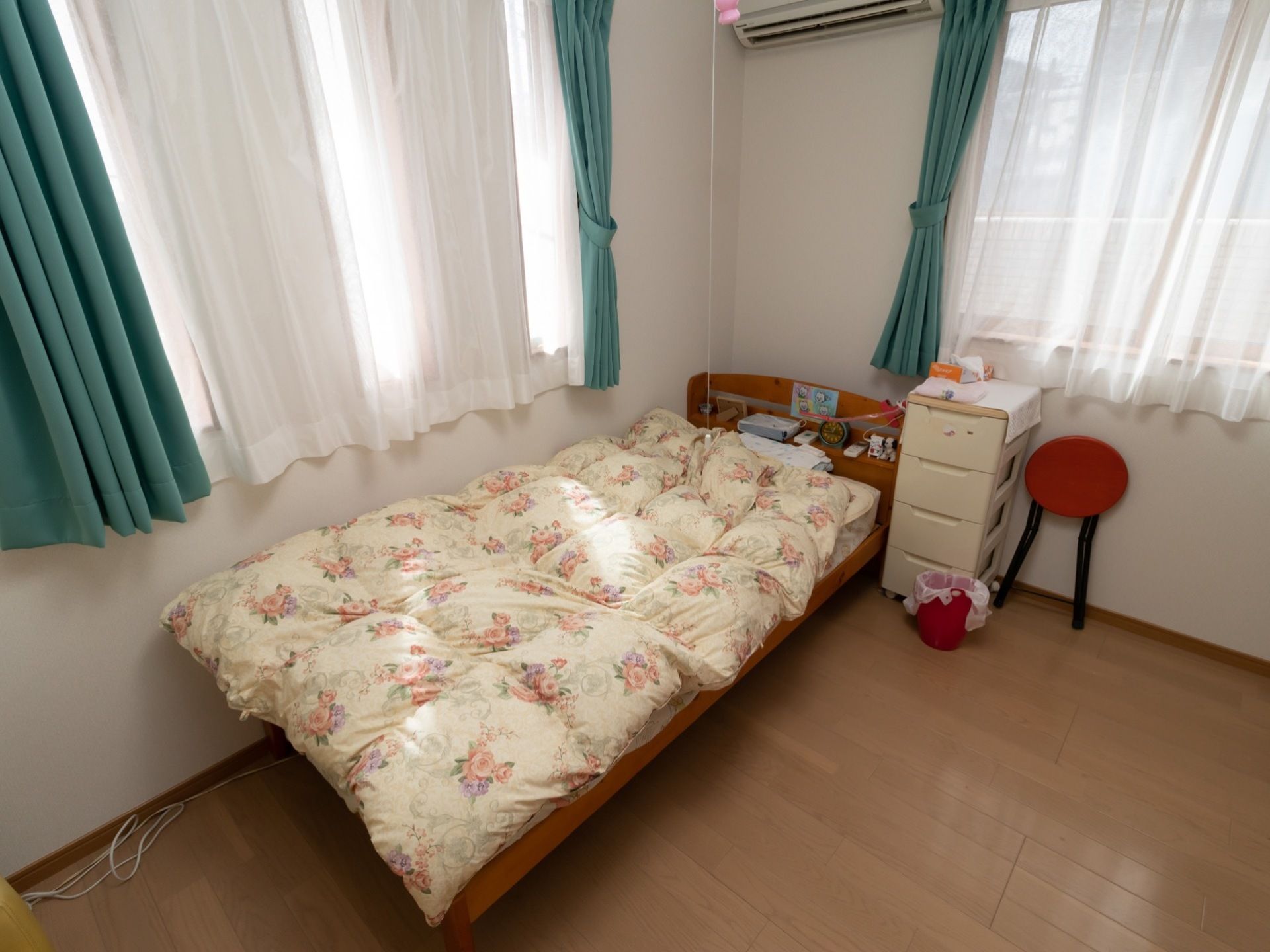 居室にはベッドとエアコンなどが設置されている。ベッドには花柄の布団が掛けられていて、横に白いチェストが置かれている。