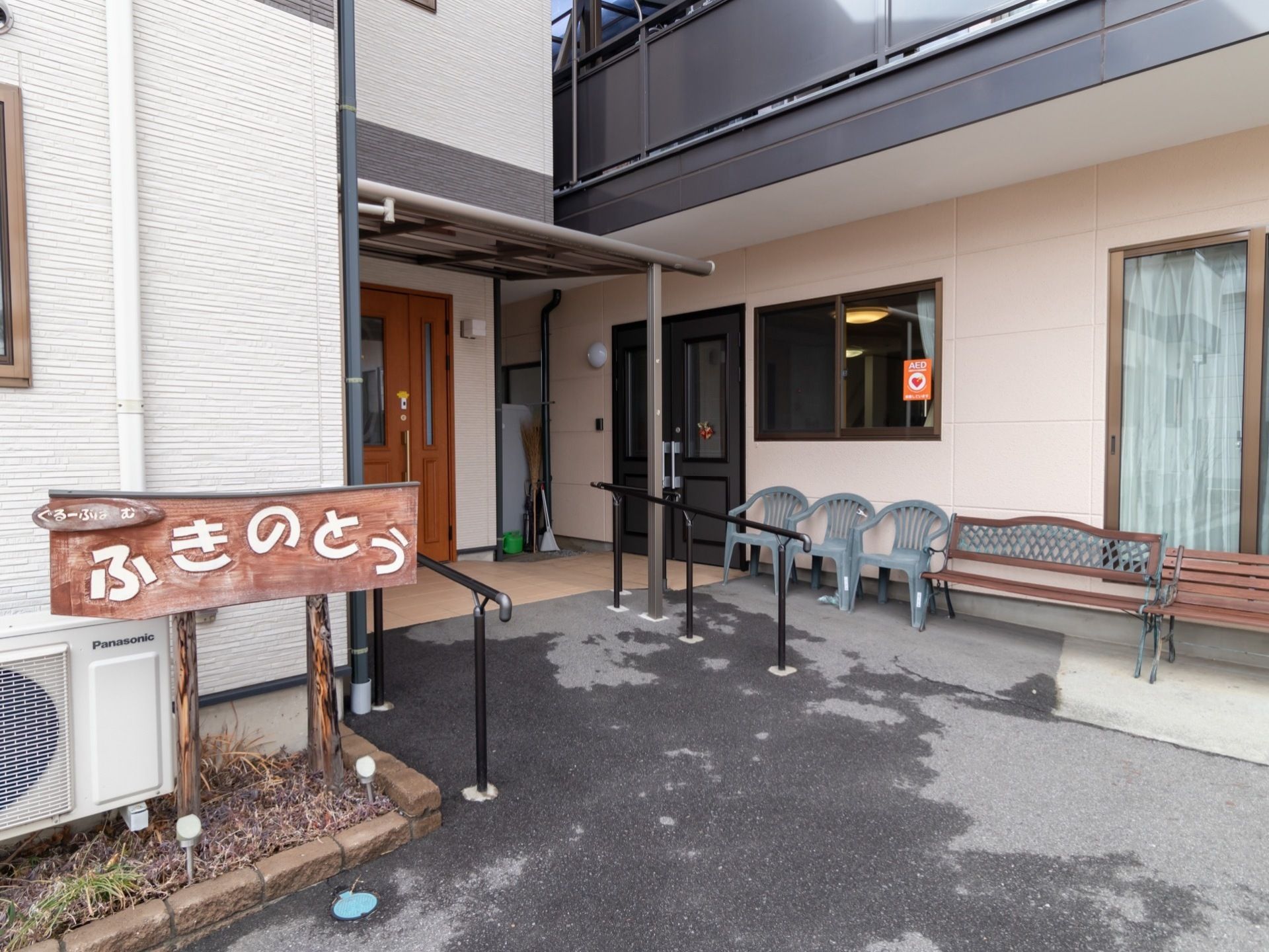 手前にある壁の前に木製の板に白い文字で施設名が書かれた看板が設置されている。玄関ドアの両サイドに黒い手すりがある。
