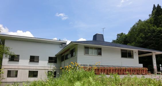 青い空の下に立っている白を基調とした建物。周りには草花が生えている。