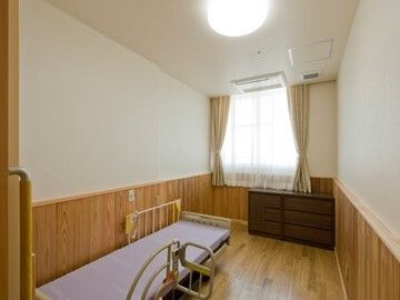 レースカーテンとカーテンが付いた窓があるフローリングの部屋には介護ベッドがあり、緊急通報装置もある。