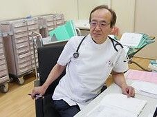 カルテの机の上に広げていて、椅子に座っている眼鏡をかけた高齢男性の医師。