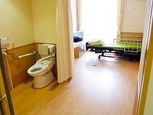 フローリングの部屋。カーテンで開け閉めするタイプのトイレや介護ベッドがあり、大きな窓が付いている。