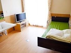 寝具付きのベッドが置かれ車いす対応の洗面所があり、テレビが設置されたフローリングの部屋。