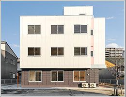 ベージュとブラウンの３階建ての建物の周りには木が植えられ、駐車スペースもある。