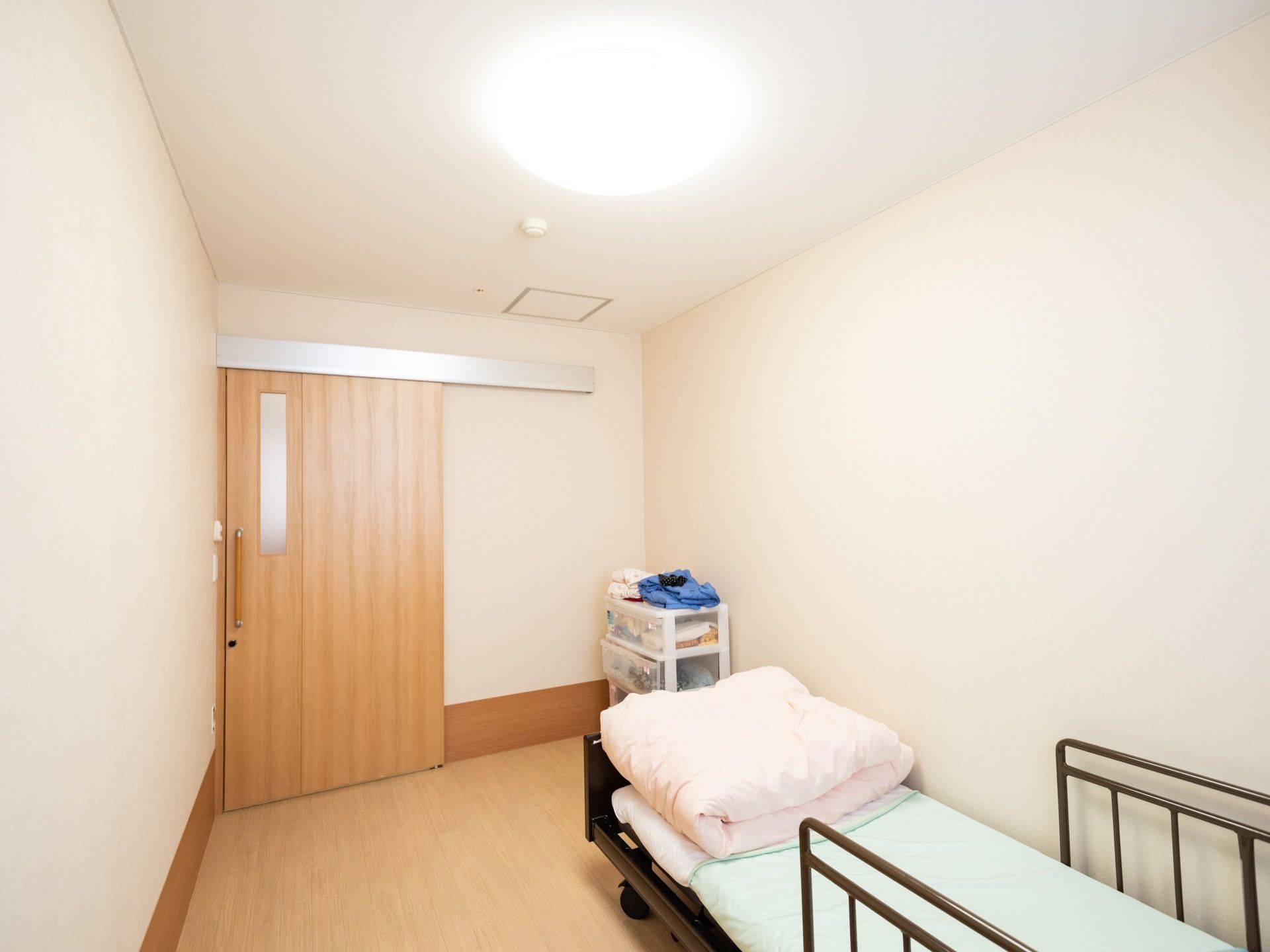 木目調の引き戸が付いた居室がある。入口近くには衣装ケースがあり、壁際には介護用ベッドが置かれている。