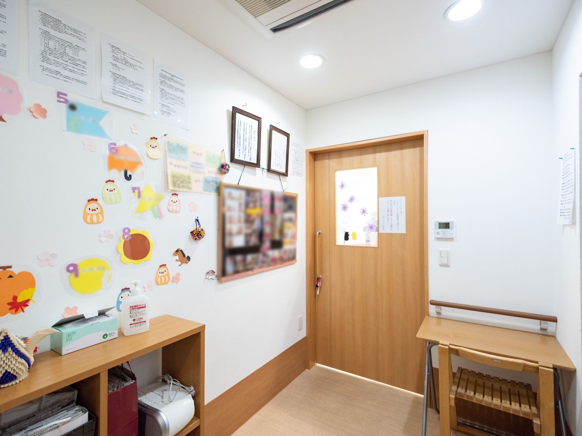 空調の付いた廊下がある。壁には写真の飾られた掲示板や、色紙の作品が貼られている。棚の上には消毒液やマスクが置かれている。