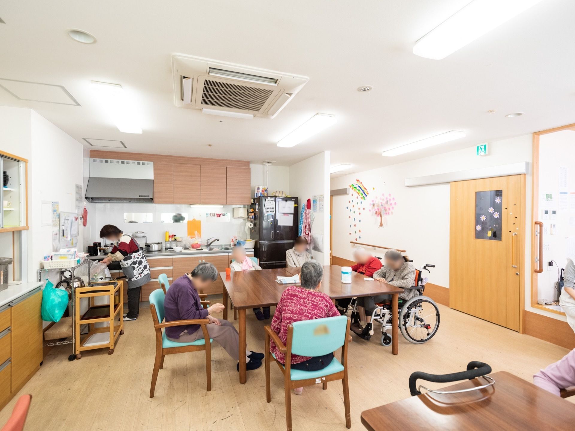 テーブルセットが並べてある食堂に入居者が集まっている。奥にはキッチンがあり、職員が食事の準備をしている。