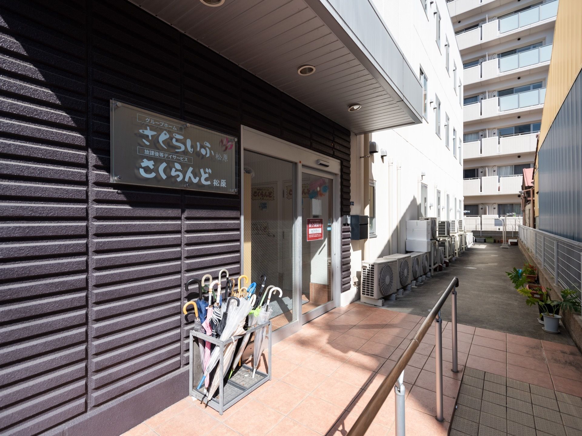 自動ドアの玄関がある。アプローチは手すりの付いたスロープになっており、ドアの横には傘立てが置かれている。