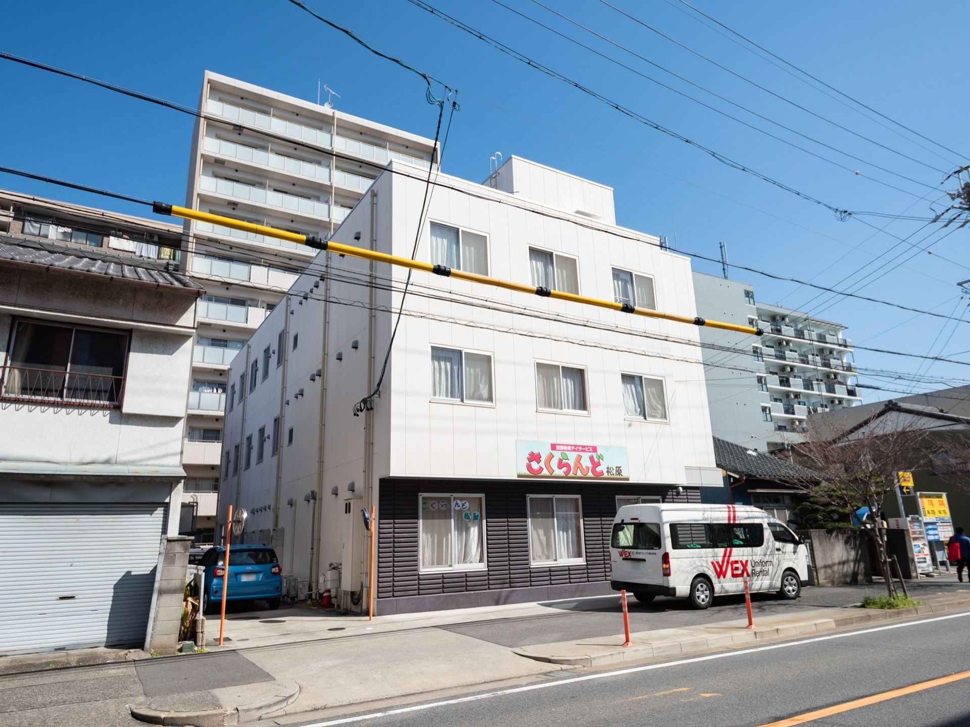 集合住宅や戸建ての多い場所に３階建ての施設がある。建物の前には幅の広い歩道があり、白いバンが１台停められている。