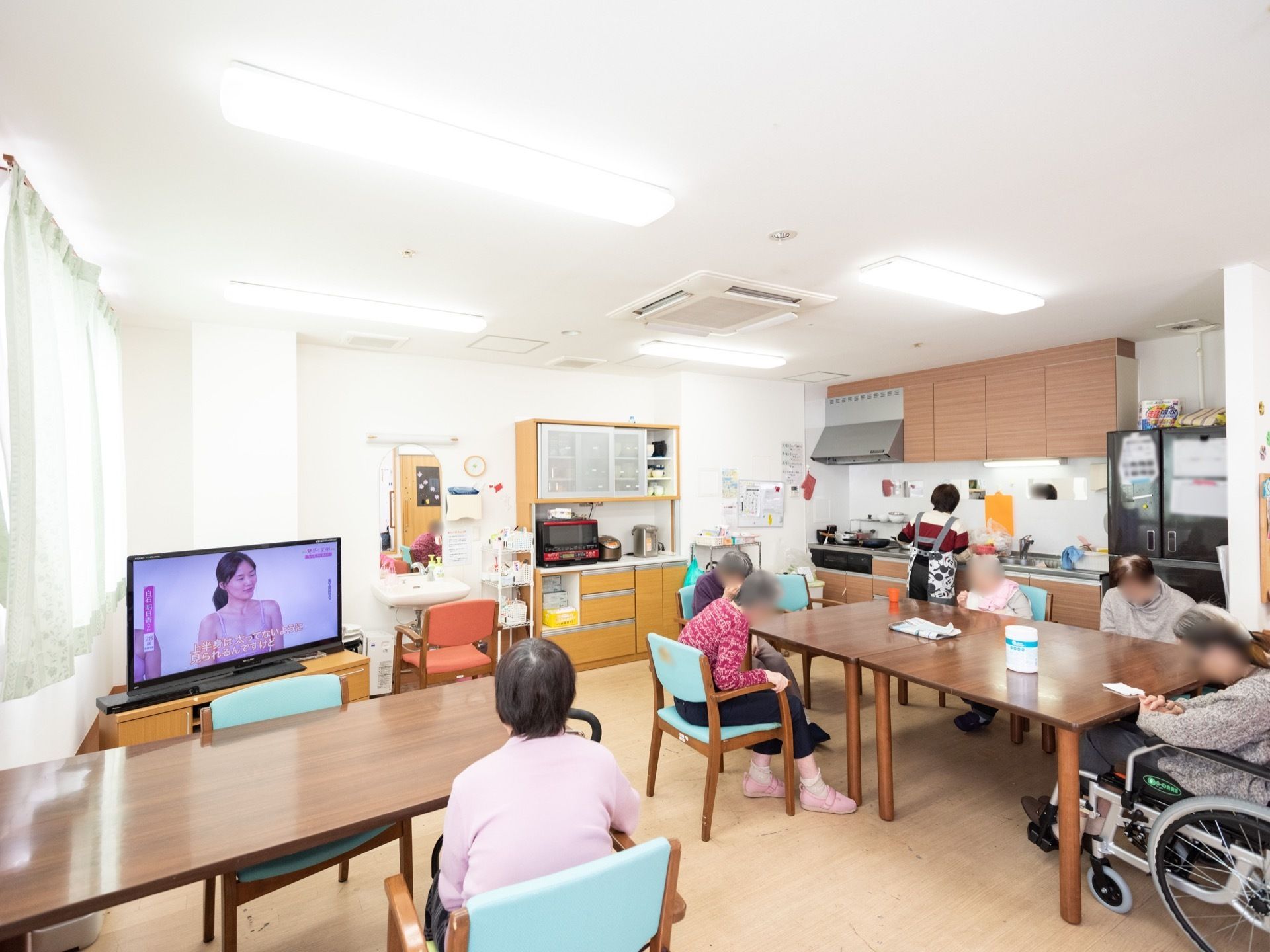 キッチンでスタッフが食事の支度をしている。テーブルには入居者が集まり、テレビ鑑賞や談笑をして過ごしている。