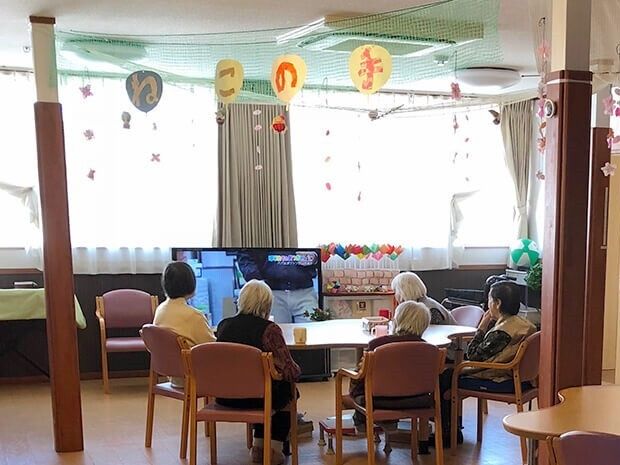 手作りの装飾品が飾られたホームの共有スペースにはテレビが置かれ、テーブルを囲んで座っている高齢者たちが見ている。