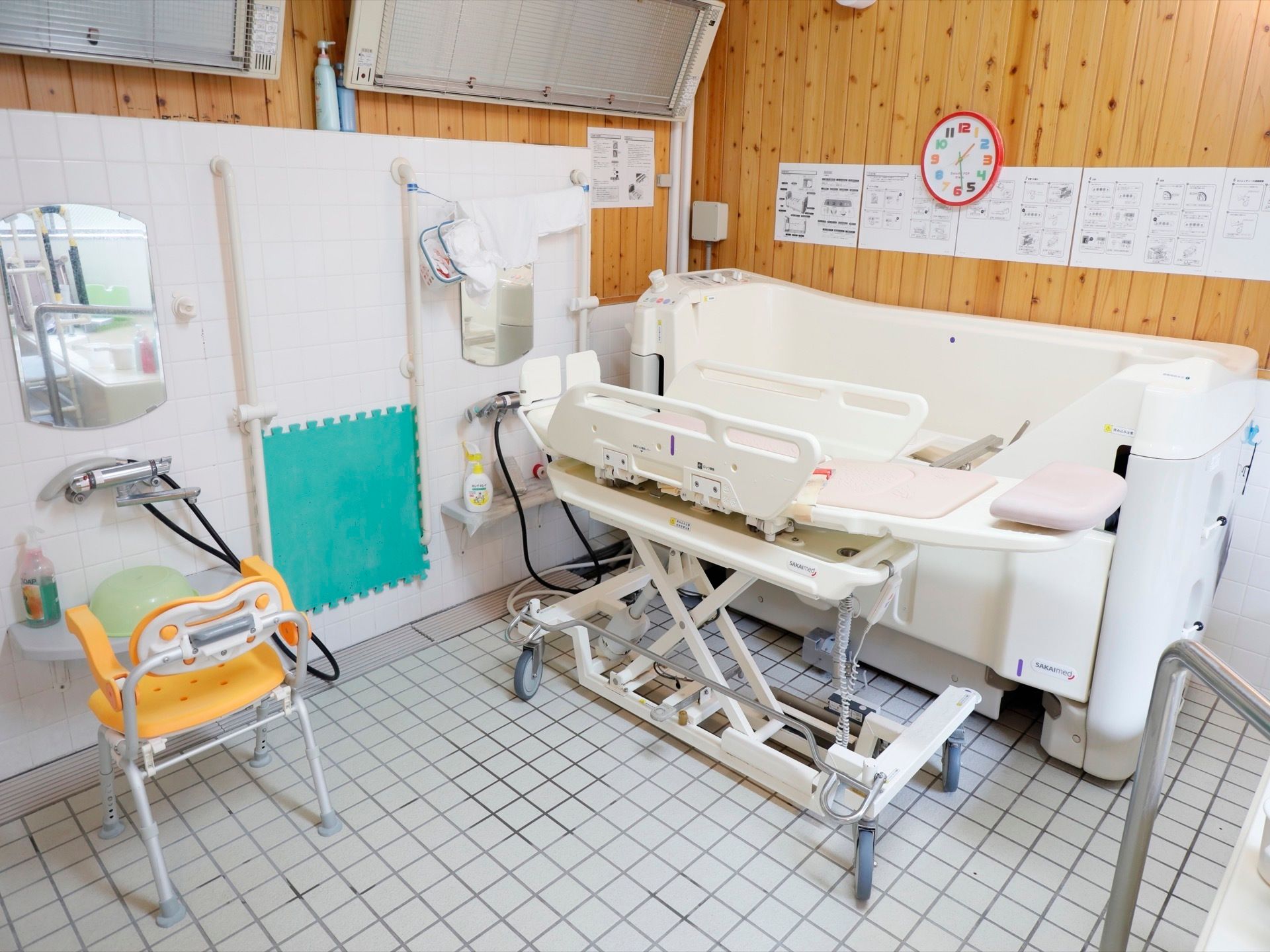介護用の入浴機械が、右側に備え付けられている。左側には洗い場があり、黄色いシャワーチェアや洗面器が置いてある。