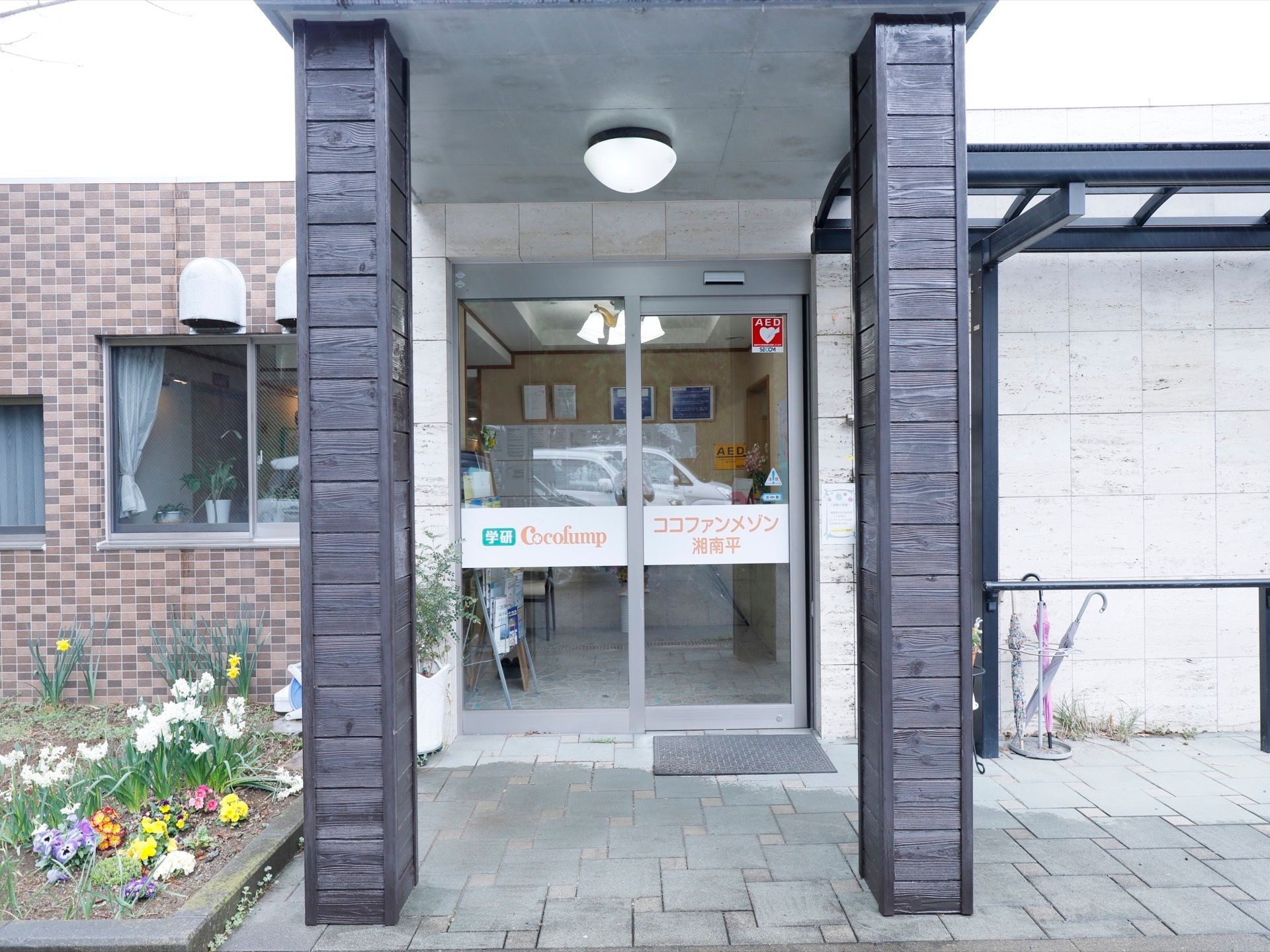 ガラス張りの自動ドアの前方には、ひさしが取り付けられている。その左横に花壇があり、パンジーや水仙が植えてある。
