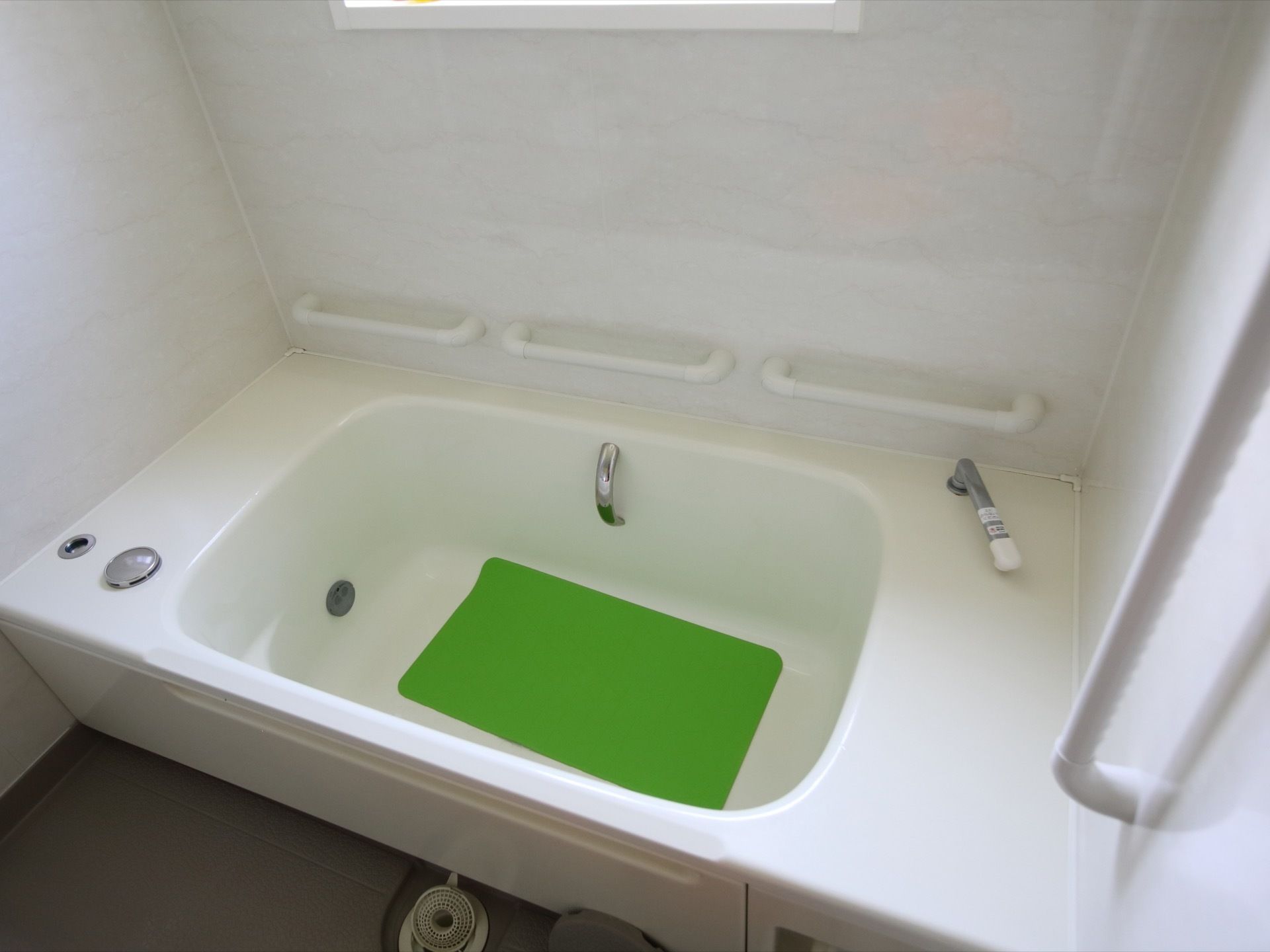 白い壁に囲まれた浴槽がある。浴槽には滑り止めマットが敷かれている。壁には窓があり、手すりが並んで３つ設置されている。