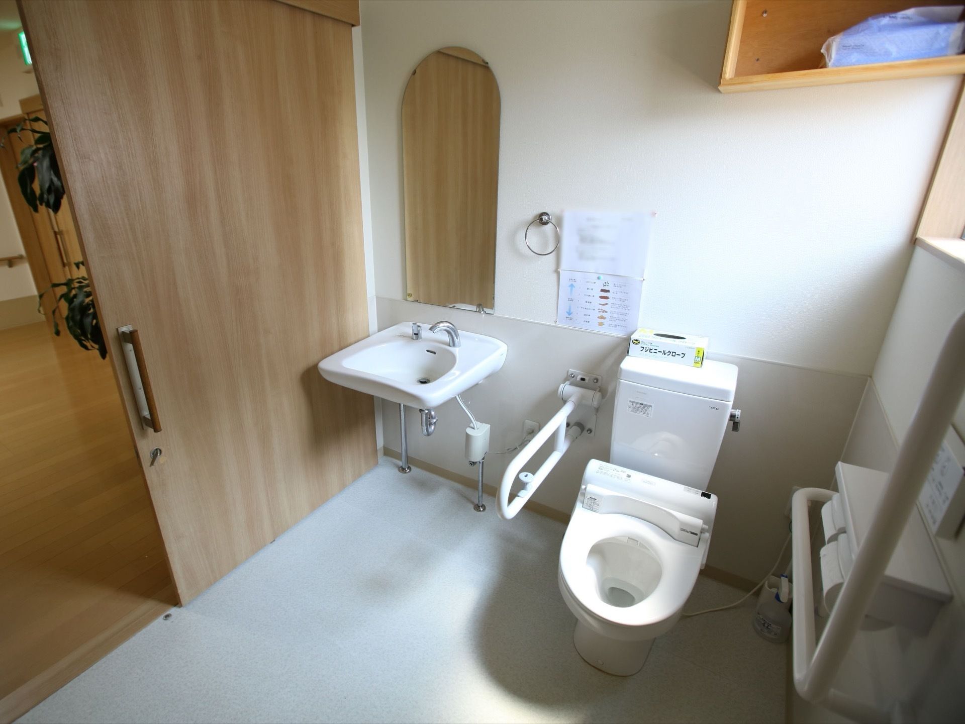 木目調のスライド式ドアが付いている広いトイレがある。鏡と洗面台が同じ空間にある。壁には手すりがあり、便座の側にひじ掛けが付いている。