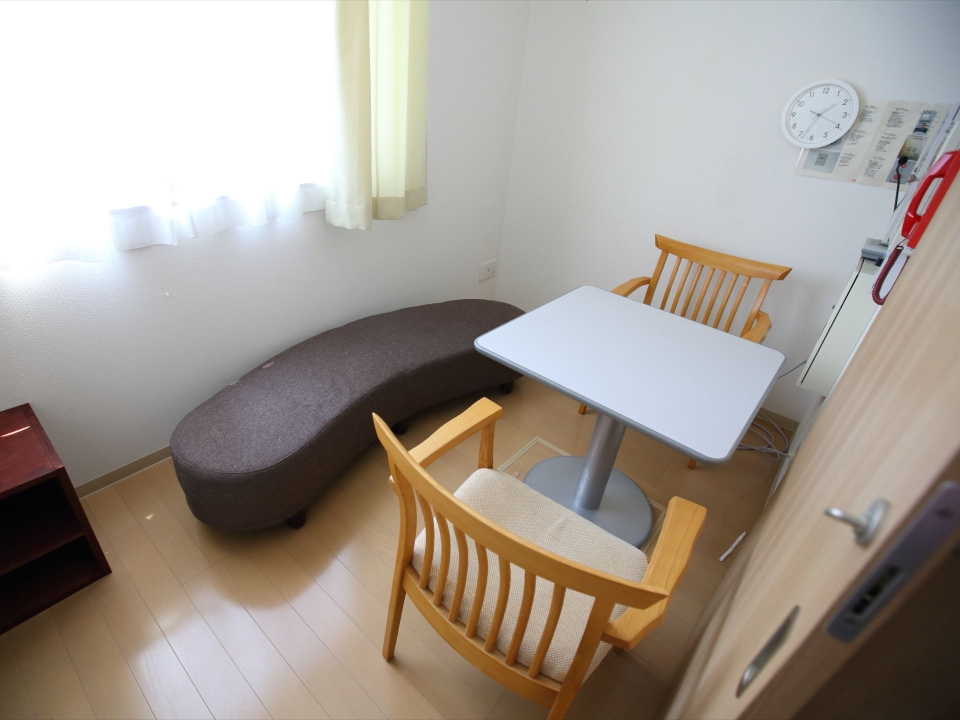 白い壁に囲まれた窓のある小部屋がある。小さいテーブルと椅子が２つあり、側にはクッション型の茶色いベンチソファが置かれている。