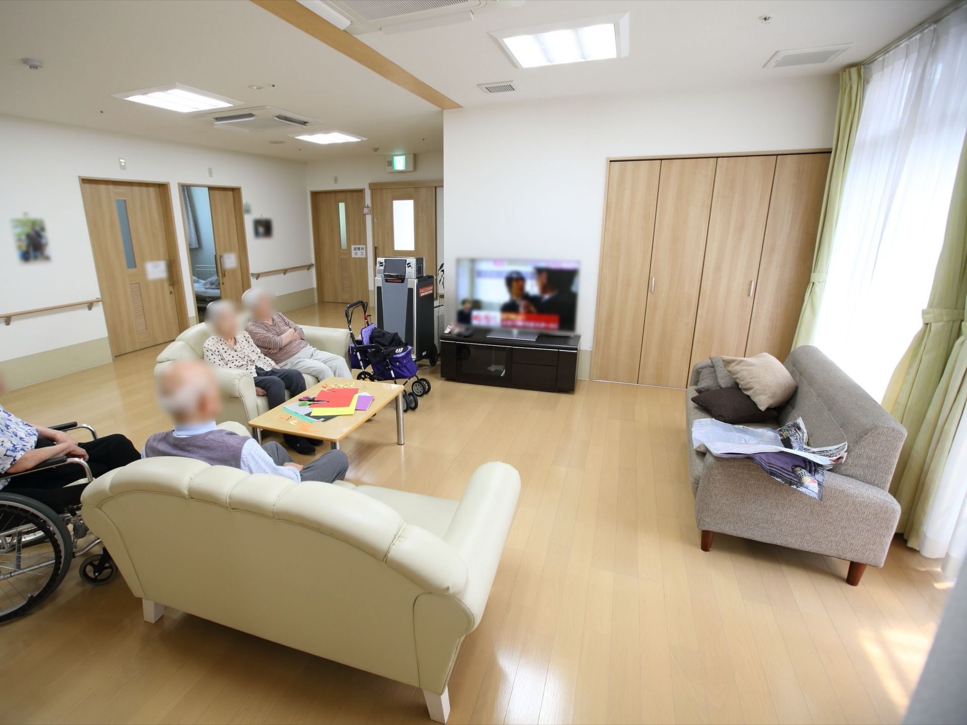 大きな窓のある談話室。ソファ、ローテーブル、テレビが置かれている。仕切りはなく、すぐ側には居室のドアが並んでいる開放的な間取りになっている。