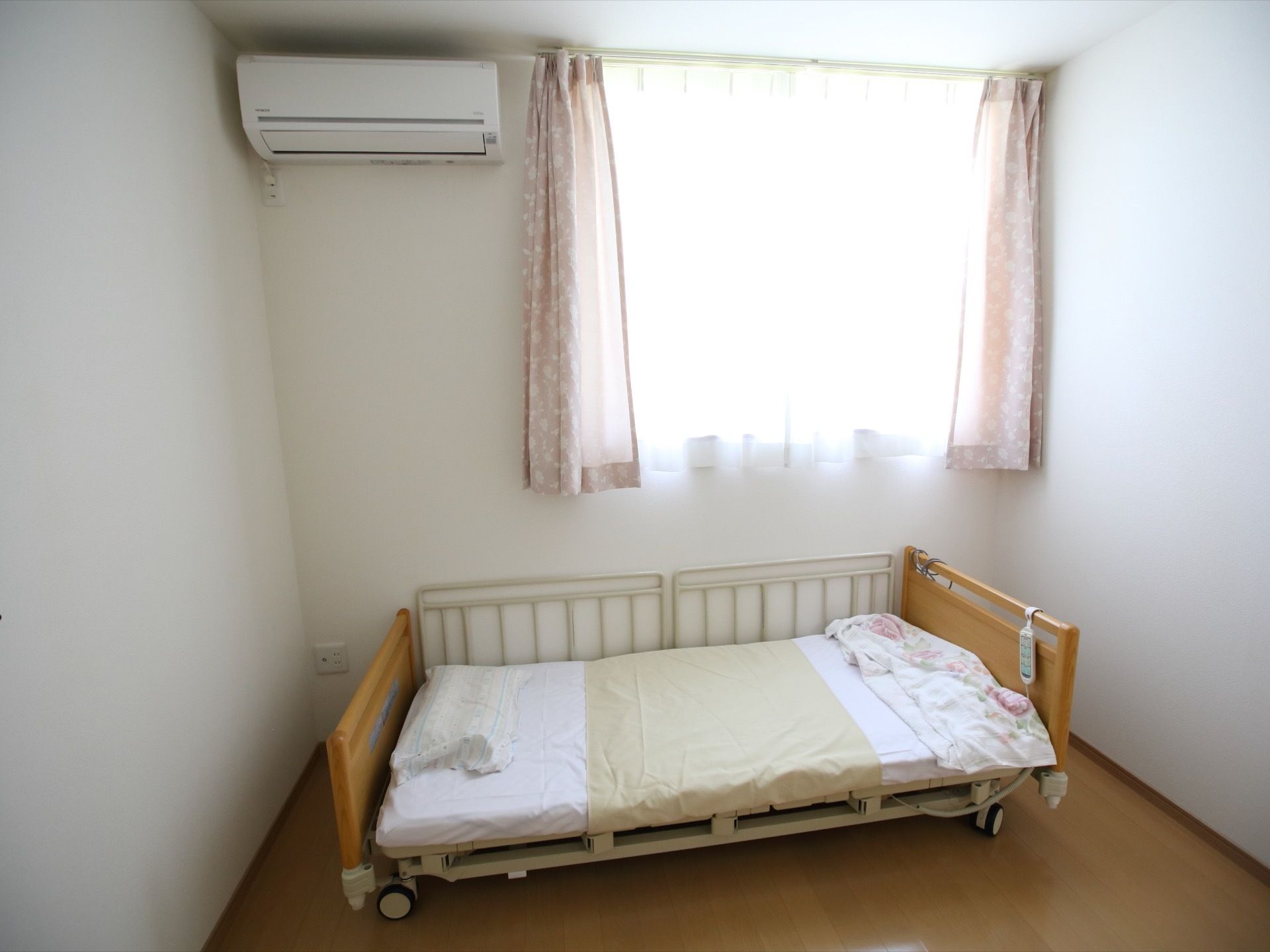 白い壁とフローリングの床の居室がある。カーテンの掛かった窓があり、側には介護用ベッドが置かれている。壁にはエアコンが設置されている。