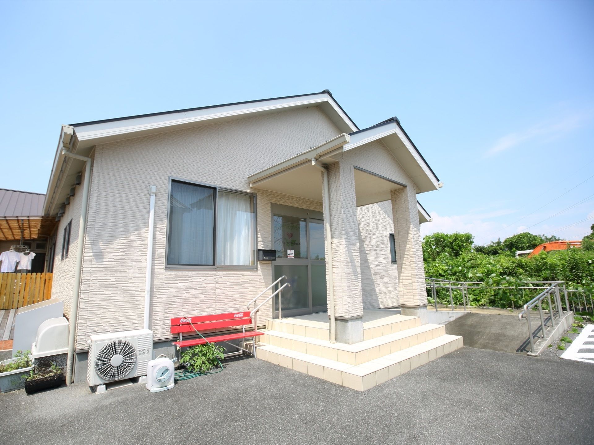 三角屋根の付いた平屋建ての建物がある。足元は舗装され、３段の階段が付いている。玄関に向かって手すりの付いたスロープが続いている。