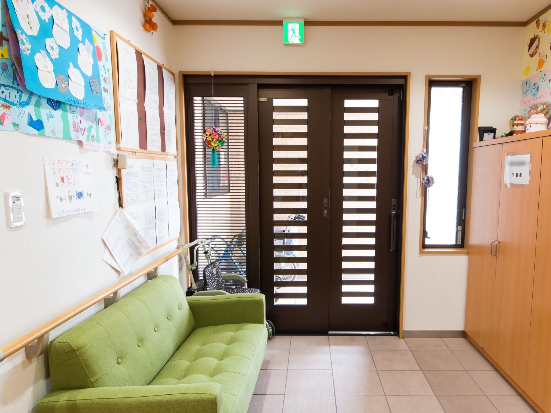 濃い茶色の引き違い扉の玄関である。天井には非常口案内灯が設置されており、壁には掲示物が複数貼られている。