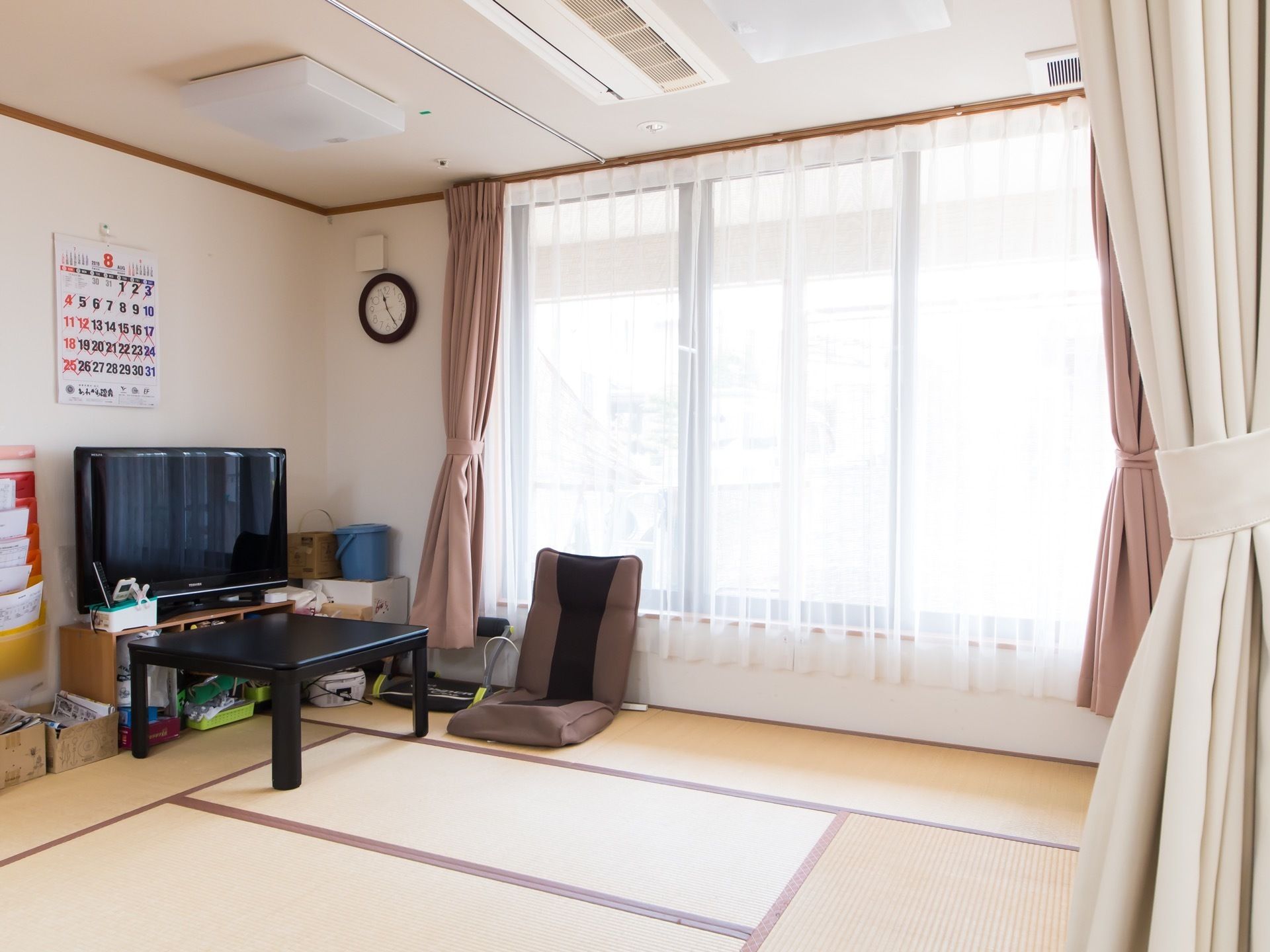 大きな窓があり、白いレースのカーテンがかけられている。天井には埋め込み式のエアコンが設置され、照明はシーリングライトとなっている。