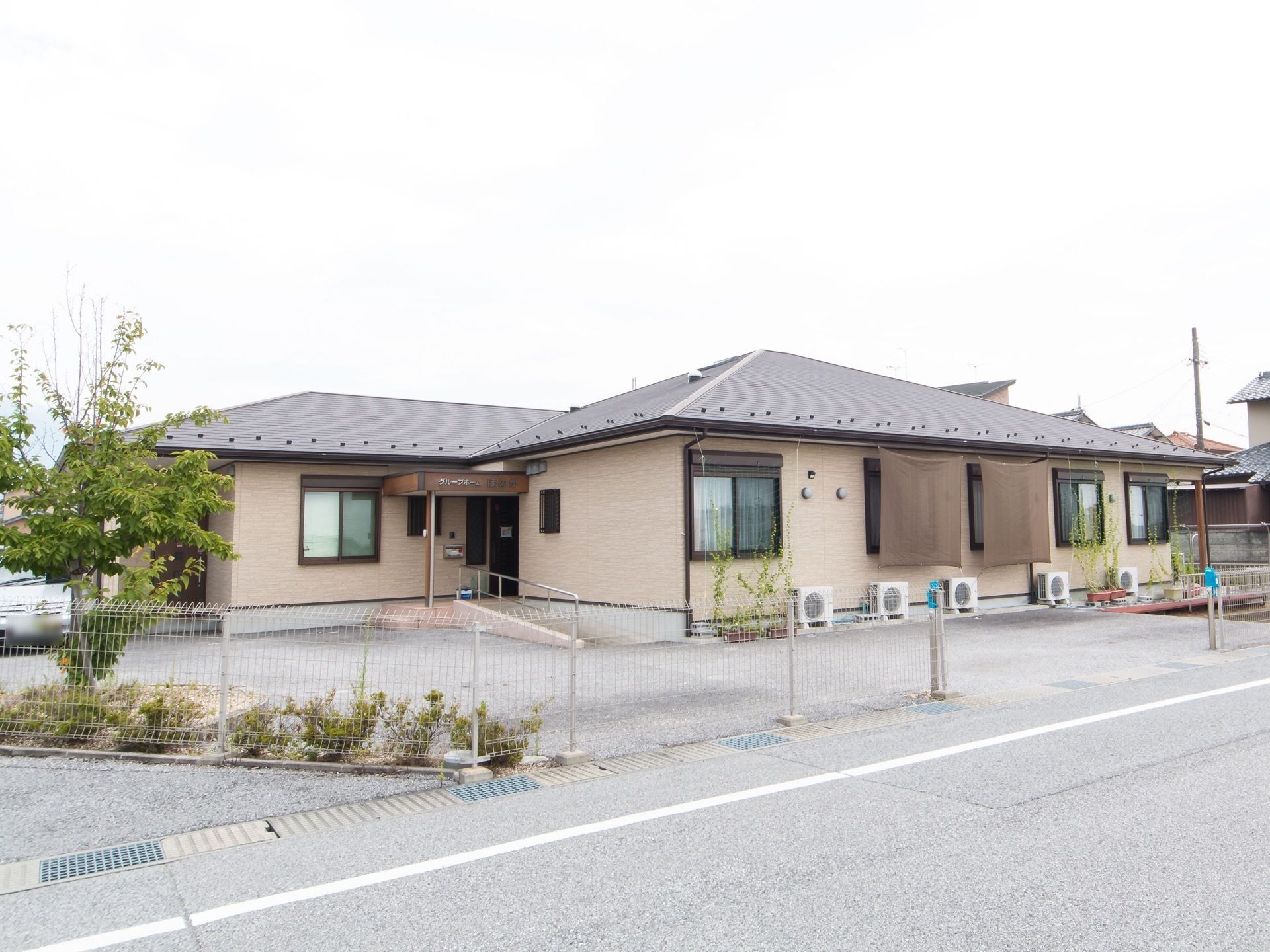 施設の敷地が、シルバーのフェンスで囲まれており、道路に面した場所である。フェンス付近には木や植物が植えられている。