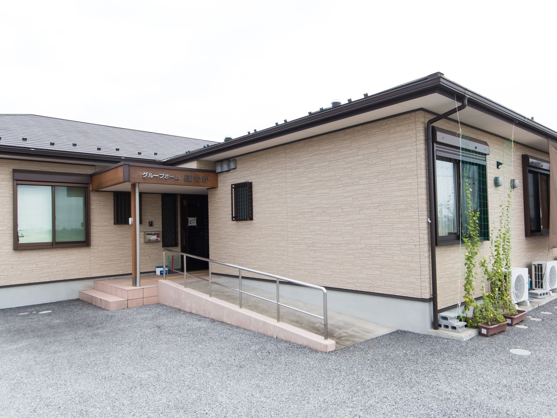 平屋建ての建物の玄関付近である。建物周囲はアスファルトで舗装されており、植物がプランターに植えてある。