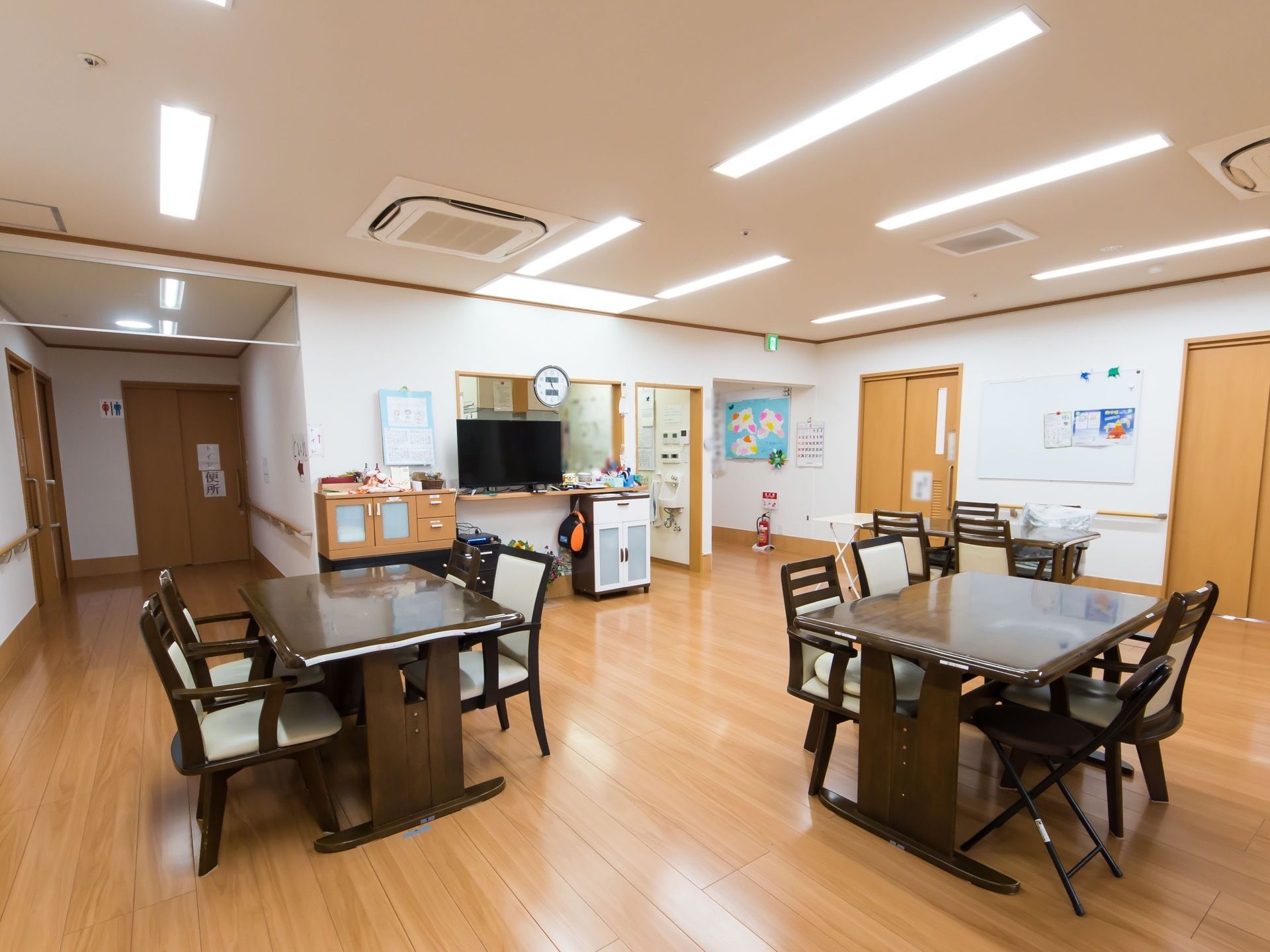 埋め込み式のエアコンが天井に設置され、細長い照明器具が複数並んでいる。フローリングの床の上にこげ茶色のテーブルが複数並べられている。