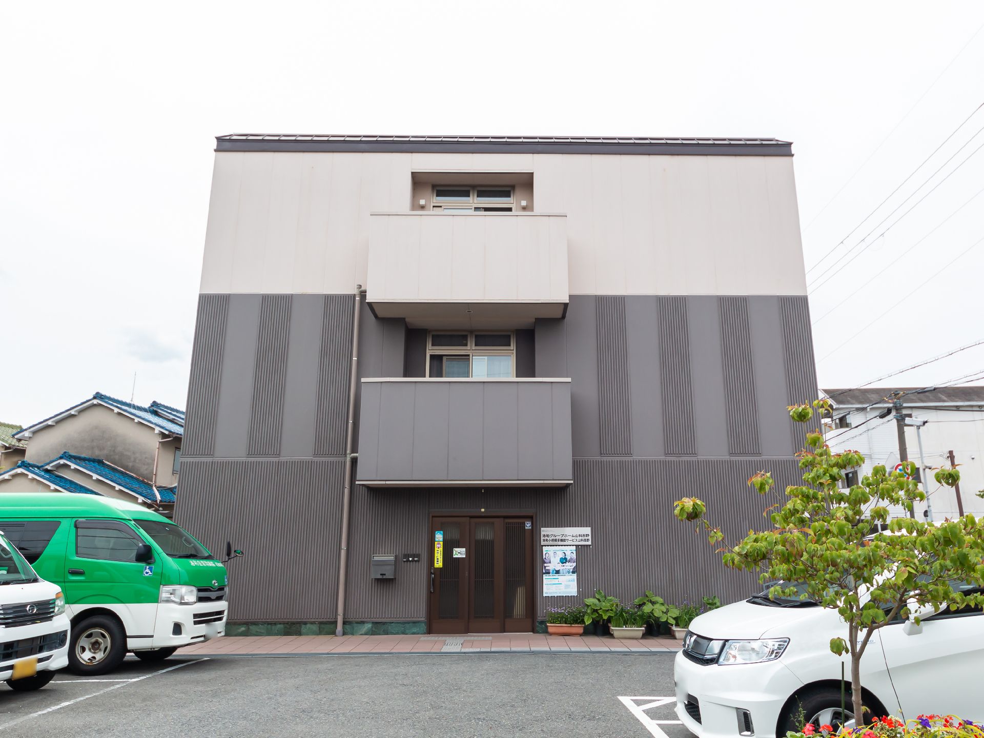 ３階建ての建物はブラウンとベージュで、駐車場の周りには花壇もある。