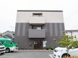 ３階建ての建物はブラウンとベージュで、駐車場の周りには花壇もある。