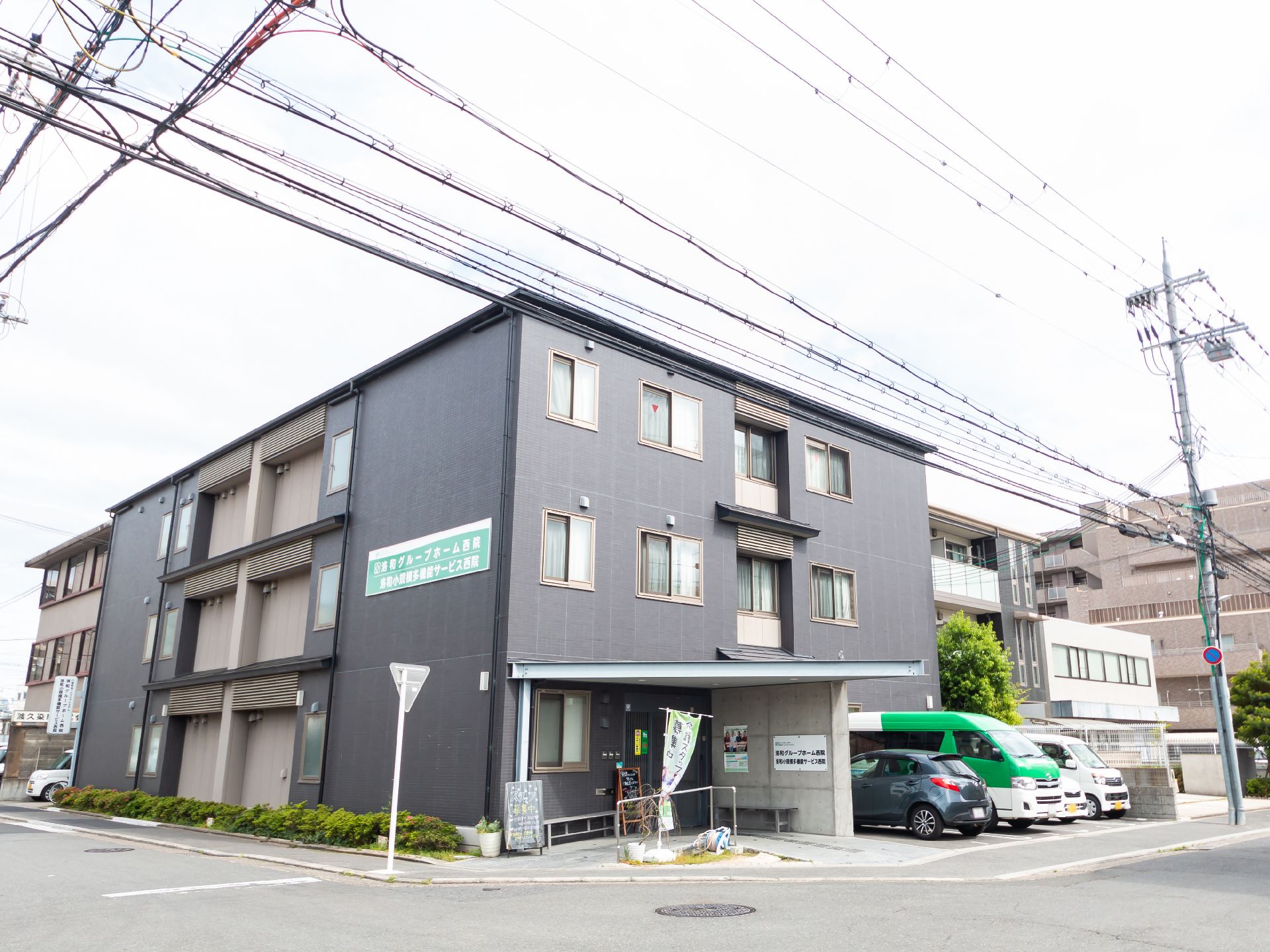 ダークグレーの3階建ての建物の玄関にそばには駐車スペースがあり、玄関には屋根が付いている。