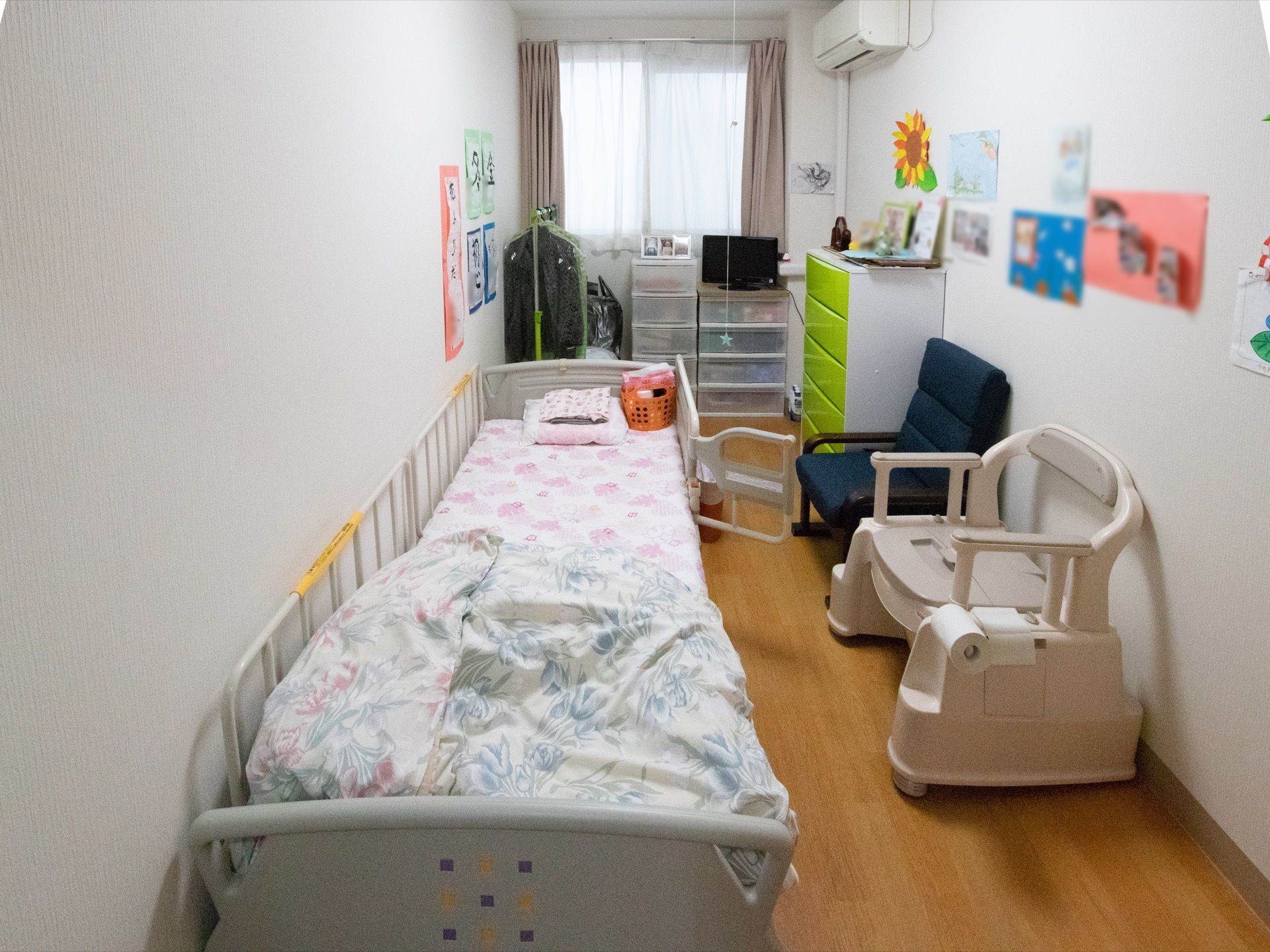 居室には、正面に窓があり整理タンスが置いてある。壁際には介護用ベッドが設置され、向い側の壁沿いには家具や椅子、ポータブル便座が置いてある。