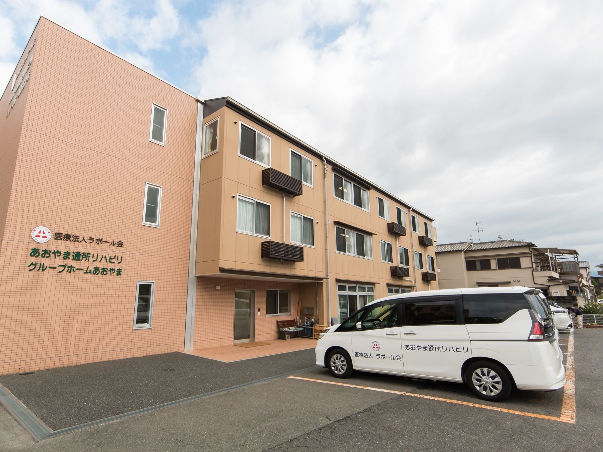 ベージュのタイルの壁に覆われている３階建ての建物がある。側面には大きく施設名が書かれており、建物の前面には駐車場がある。