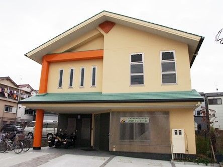ベージュを基調とした建物に三角の屋根。家庭的な雰囲気の2階建ての建物の周辺には緑もある。