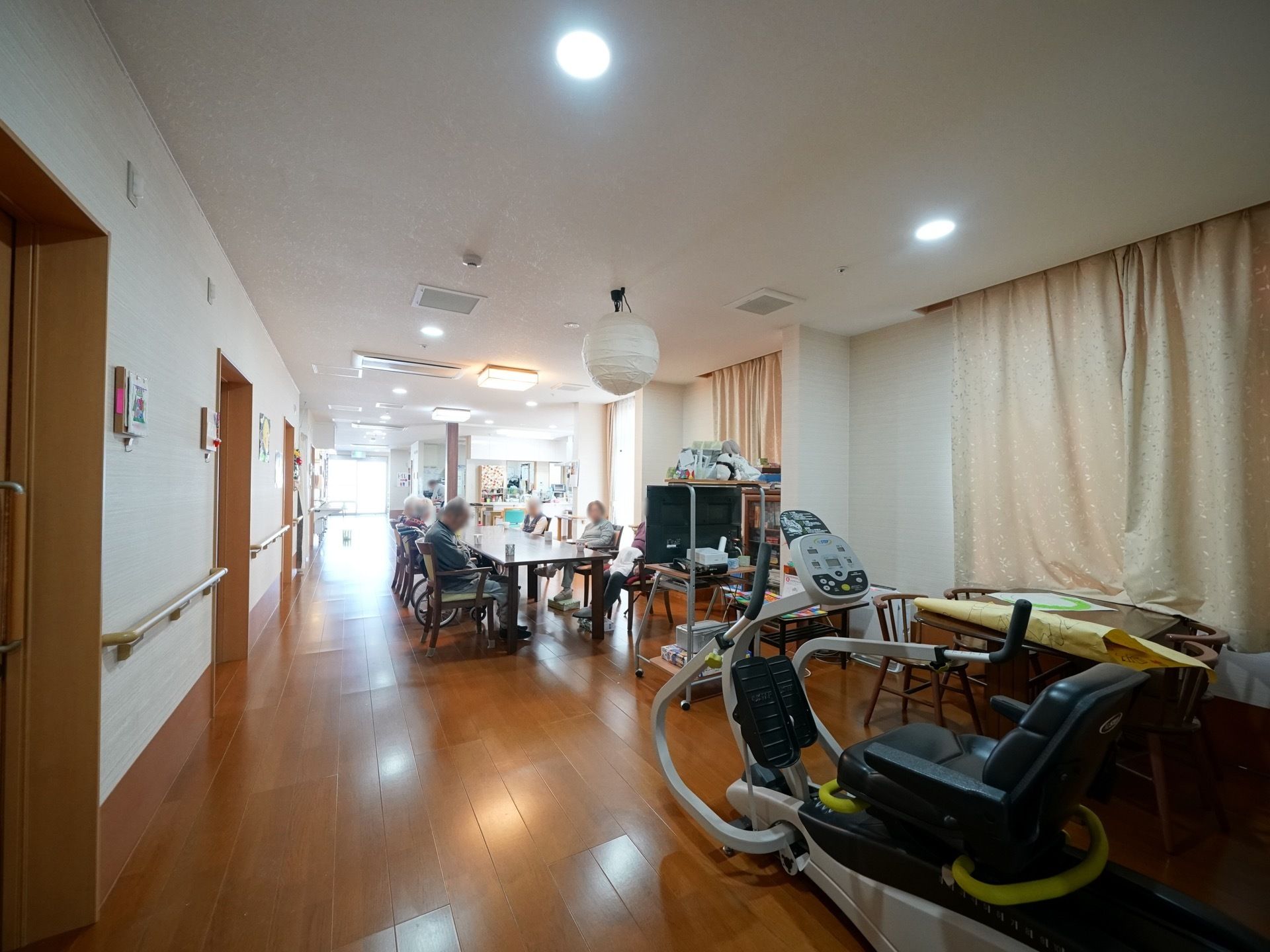 広い一室で、入居者様が食卓を囲むように談話を楽しんでいる。テレビやトレーニング用の機器も設置されている。