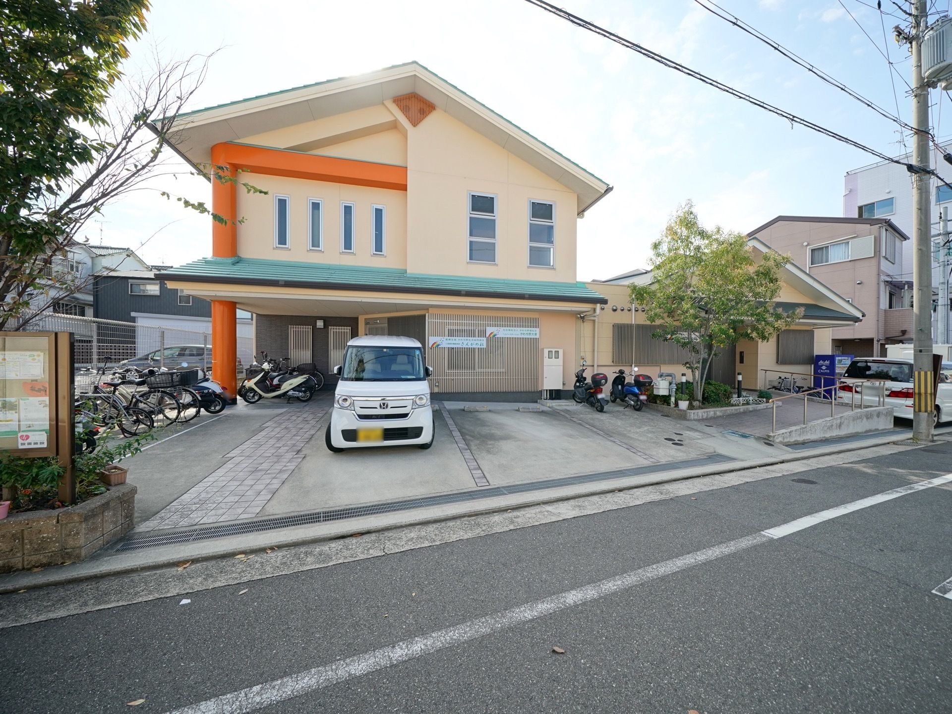 玄関前には広々とした駐車場や駐輪場がある。スロープと手すりが設置されている。建物は優しいクリームを基調とした色合いになっている。